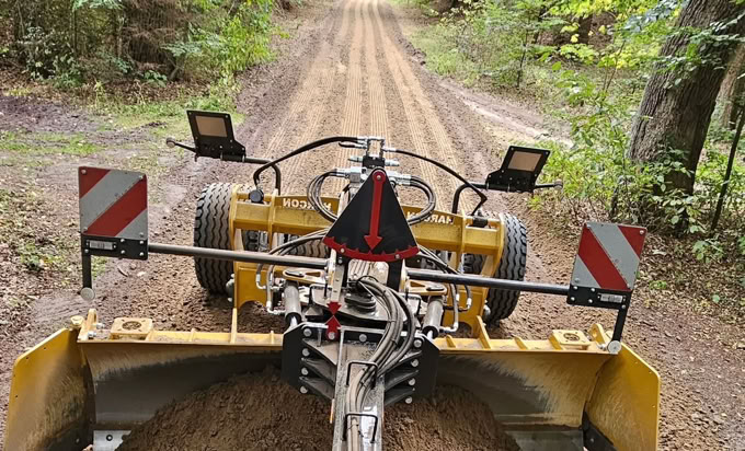 Straßenbaumaschine auf einem unbefestigten Waldweg, umgeben von Bäumen.