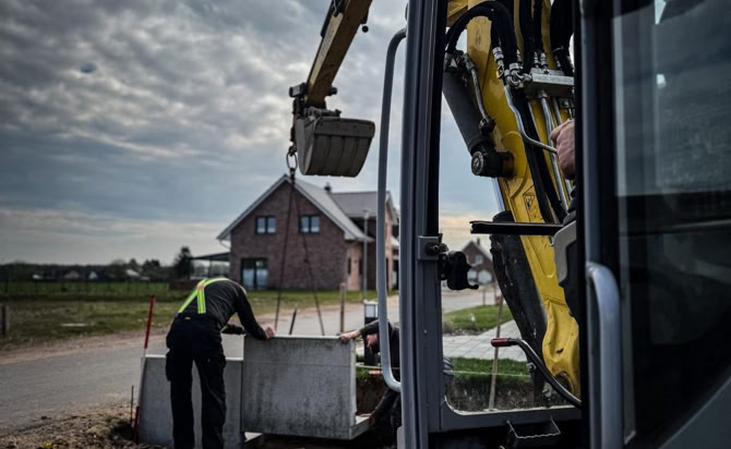 Ein Bagger hebt Betonblöcke auf einer Baustelle an, während eine Person daneben steht; im Hintergrund ist ein Haus zu sehen.