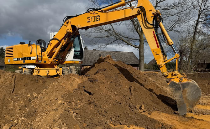 Ein gelber Bagger hebt Erde auf einer Baustelle aus, umgeben von Bäumen und einem bewölkten Himmel.
